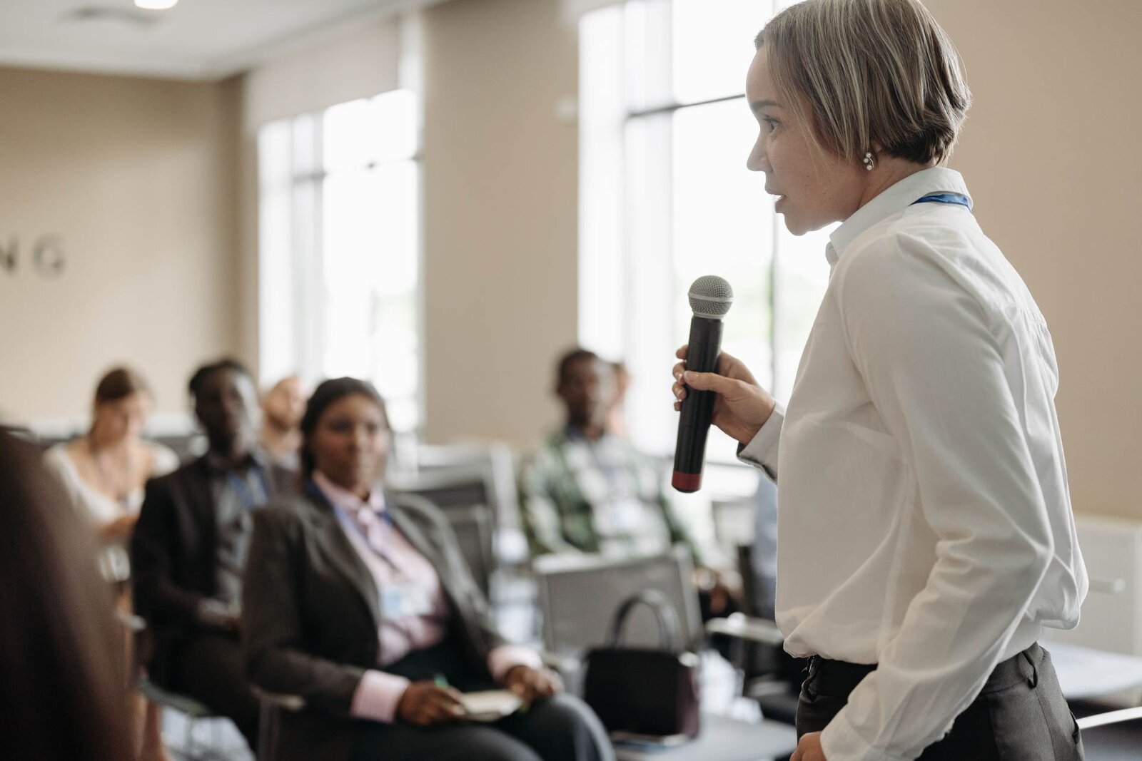 A presenter in a white shirt speaks into a handheld microphone, seen in profile, with delegates listening behind her in a bright, window-lit room