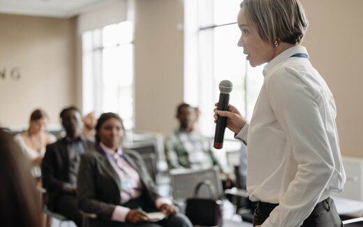 A presenter speaks into a handheld microphone before a seated audience in a bright conference room