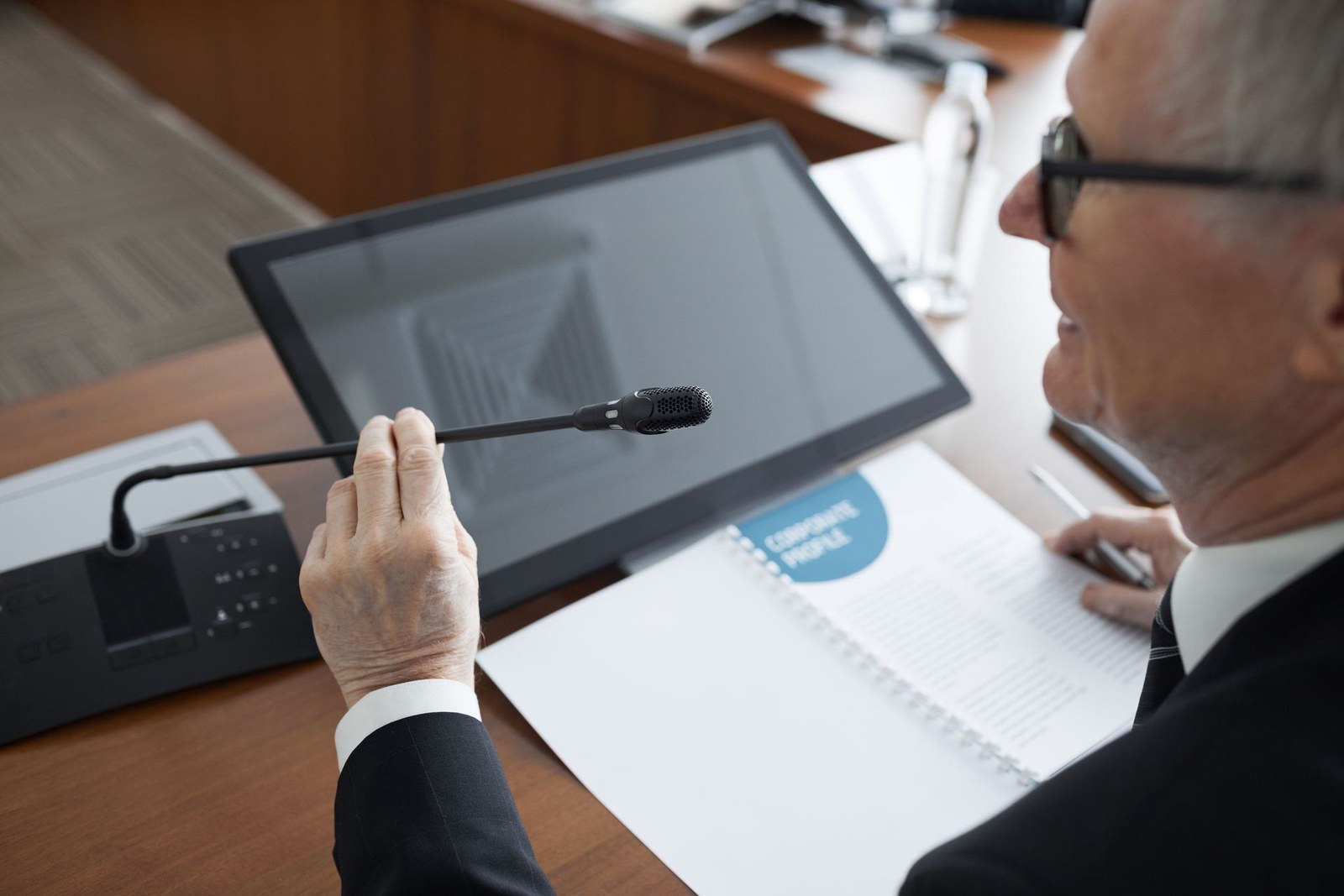 A presenter's hand adjusts a gooseneck microphone beside a monitor and briefing binder during a formal session