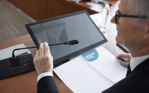 A presenter's hand adjusts a gooseneck microphone beside a monitor and briefing binder during a formal session