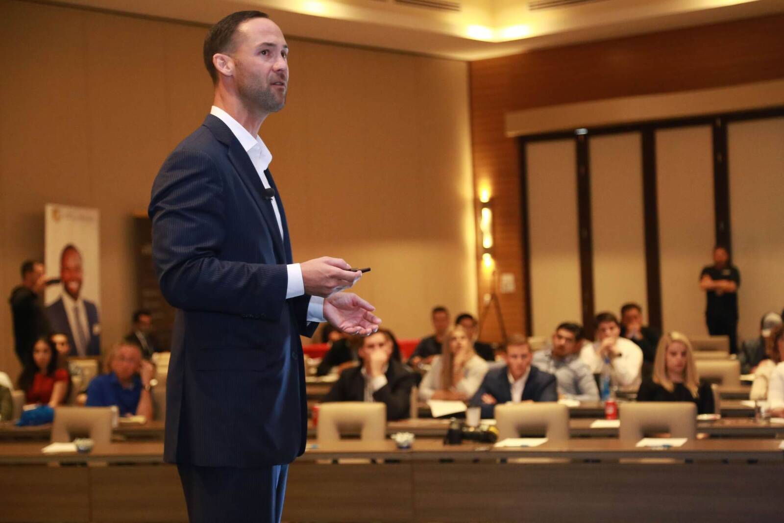 A speaker in a navy suit addresses a seated audience in a hotel conference ballroom