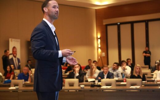 A speaker in a navy suit addresses a seated audience in a hotel conference ballroom