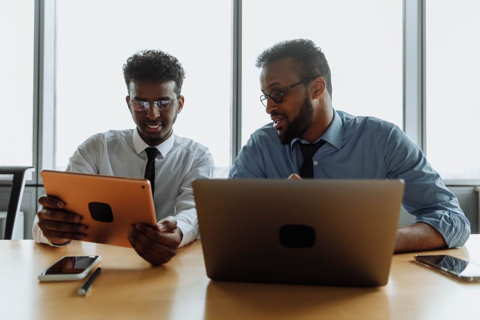 Two colleagues lean over a tablet and laptop together, discussing what is on the screen