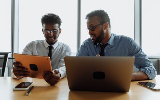 Two colleagues lean over a tablet and laptop together, discussing what is on the screen