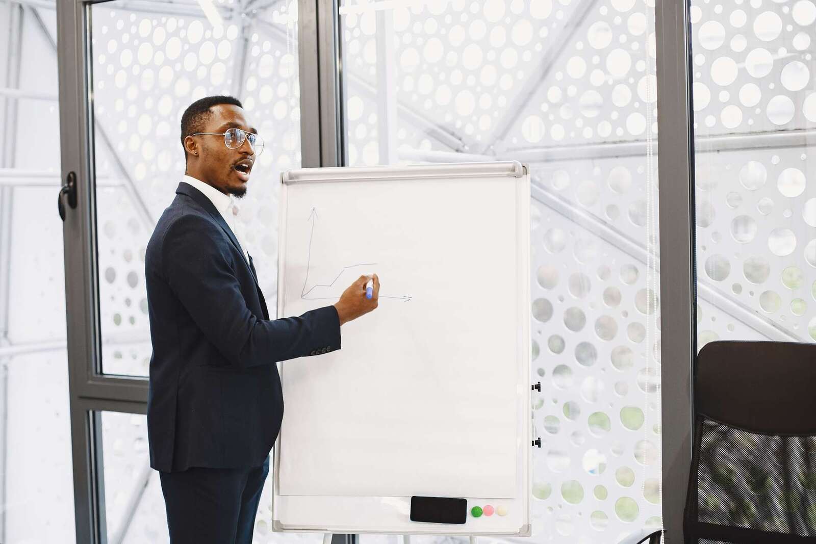 A presenter in a suit addresses a session from beside a flipchart in a bright, glass-walled venue