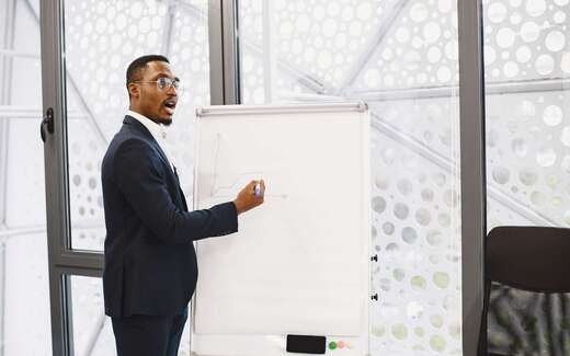 A presenter in a suit addresses a session from beside a flipchart in a bright, glass-walled venue