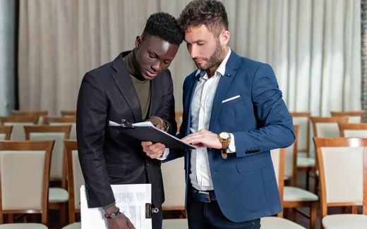 Two conference delegates reviewing a program agenda among rows of seminar chairs