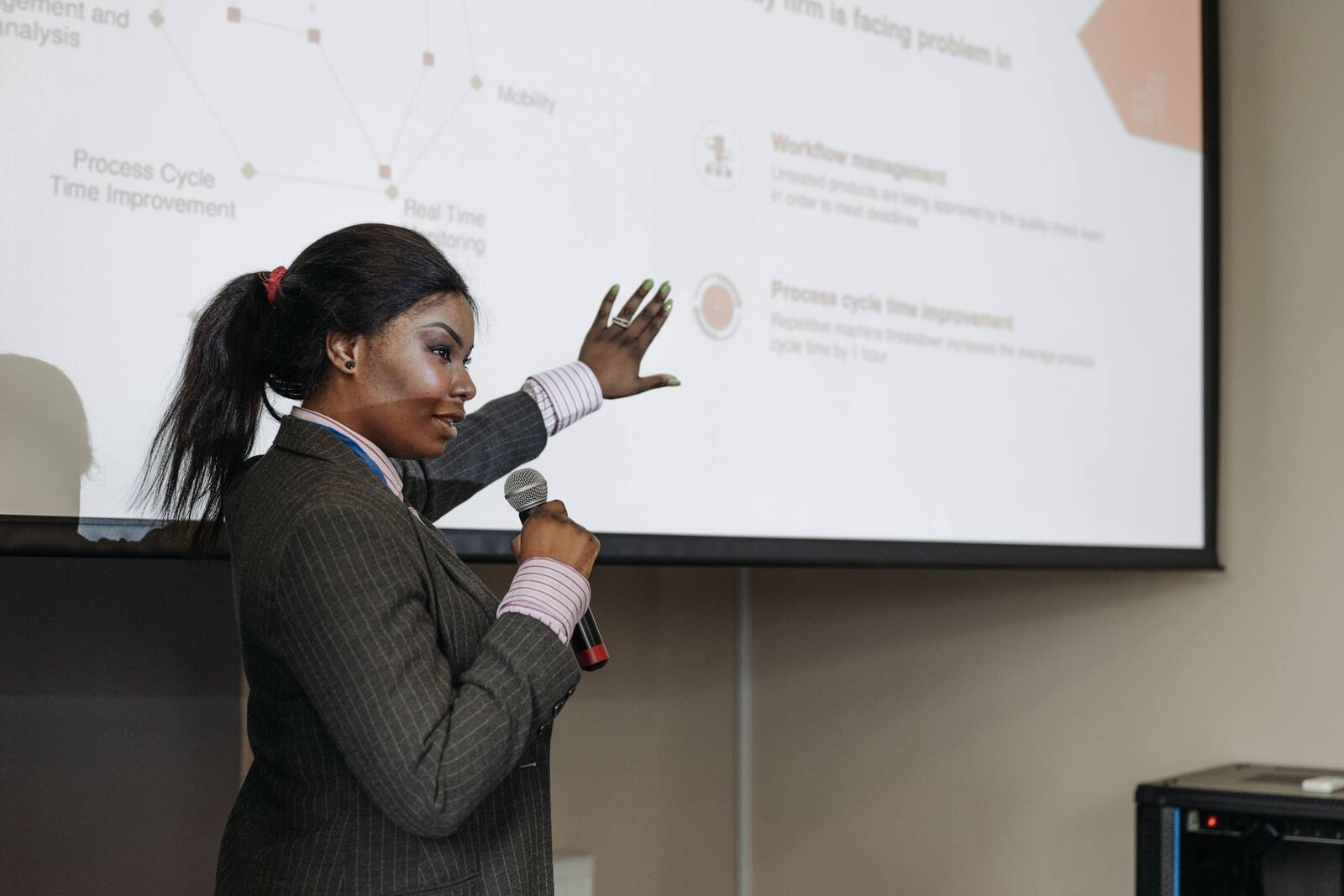 A speaker in a pinstripe blazer gestures toward a presentation slide while addressing a conference audience