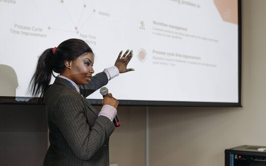 A speaker in a pinstripe blazer gestures toward a presentation slide while addressing a conference audience