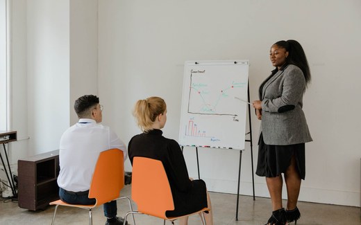 A presenter gestures toward a flipchart diagram while addressing two seated colleagues in a bright training room