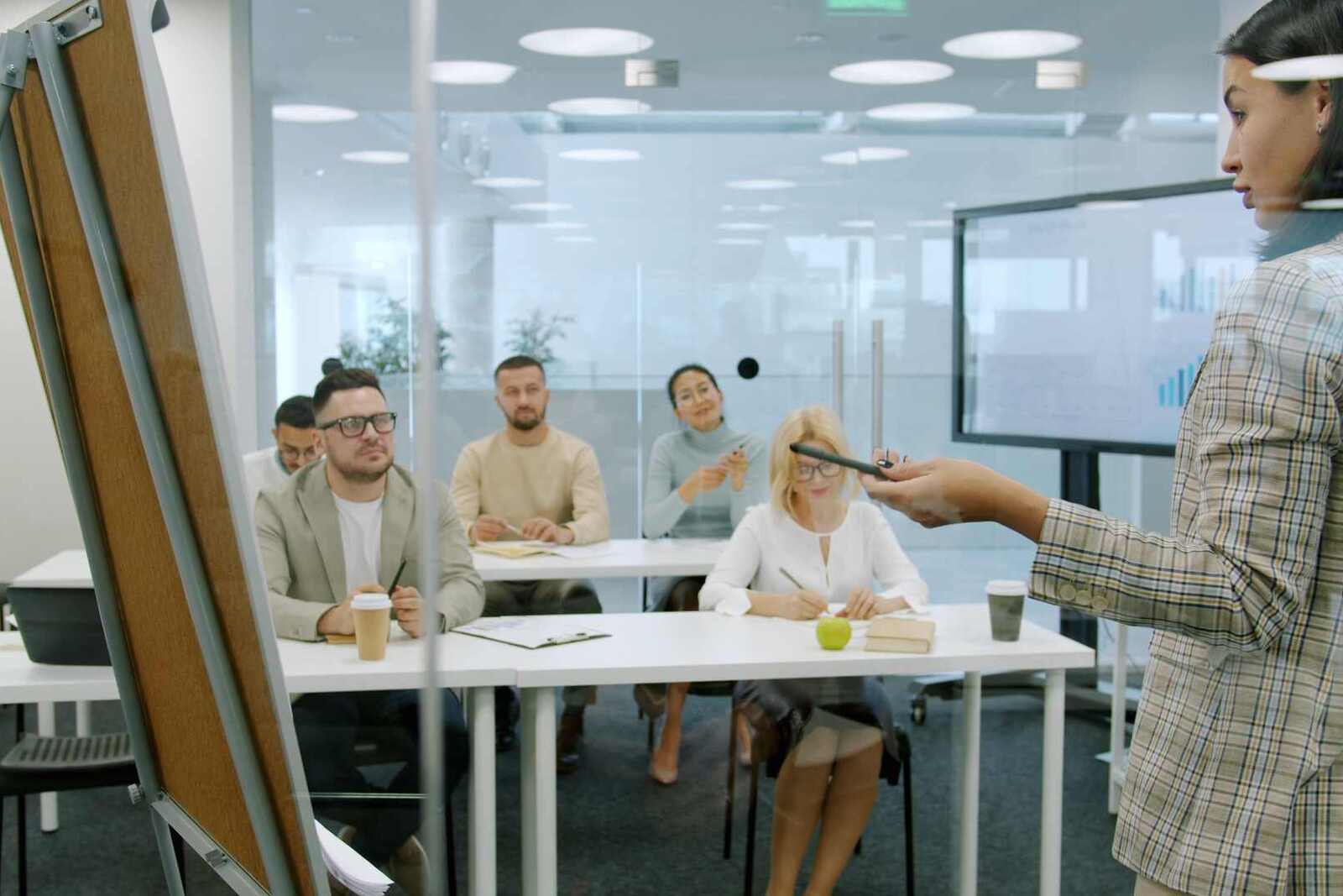 A presenter gestures toward five seated delegates taking notes in a bright, glass-walled training room