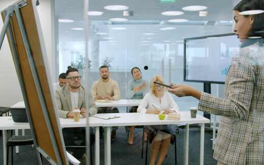 A presenter gestures toward five seated delegates taking notes in a bright, glass-walled training room