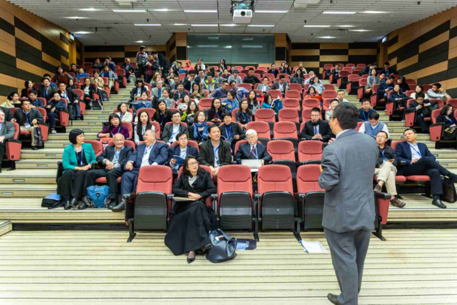 A speaker seen from behind addresses a full lecture hall of seated delegates with lanyards