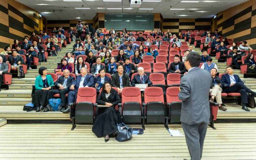 A speaker seen from behind addresses a full lecture hall of seated delegates with lanyards