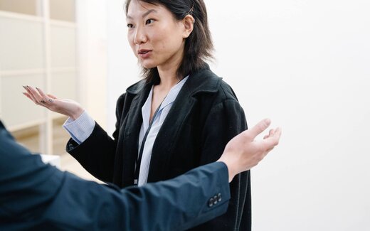 A businesswoman in a blazer, wearing a lanyard, gestures while speaking to colleagues
