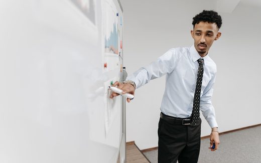 A presenter in a dress shirt and tie pointing to a chart drawn on a whiteboard