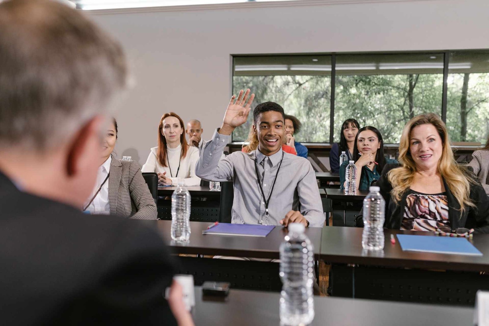 A workshop attendee raises his hand to ask a question in a bright seminar room, lanyard visible, other attendees seated around him