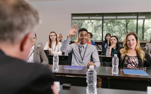 A workshop attendee raises his hand to ask a question in a bright seminar room, lanyard visible, other attendees seated around him