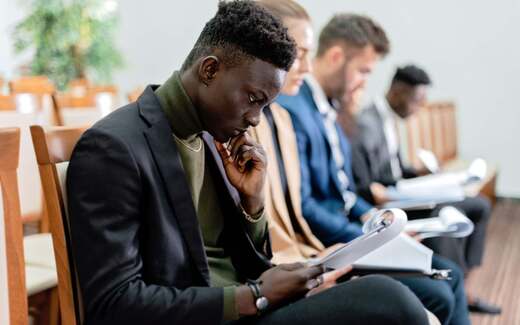 A delegate reads through papers at a seated session, chin resting on hand, other attendees out of focus alongside