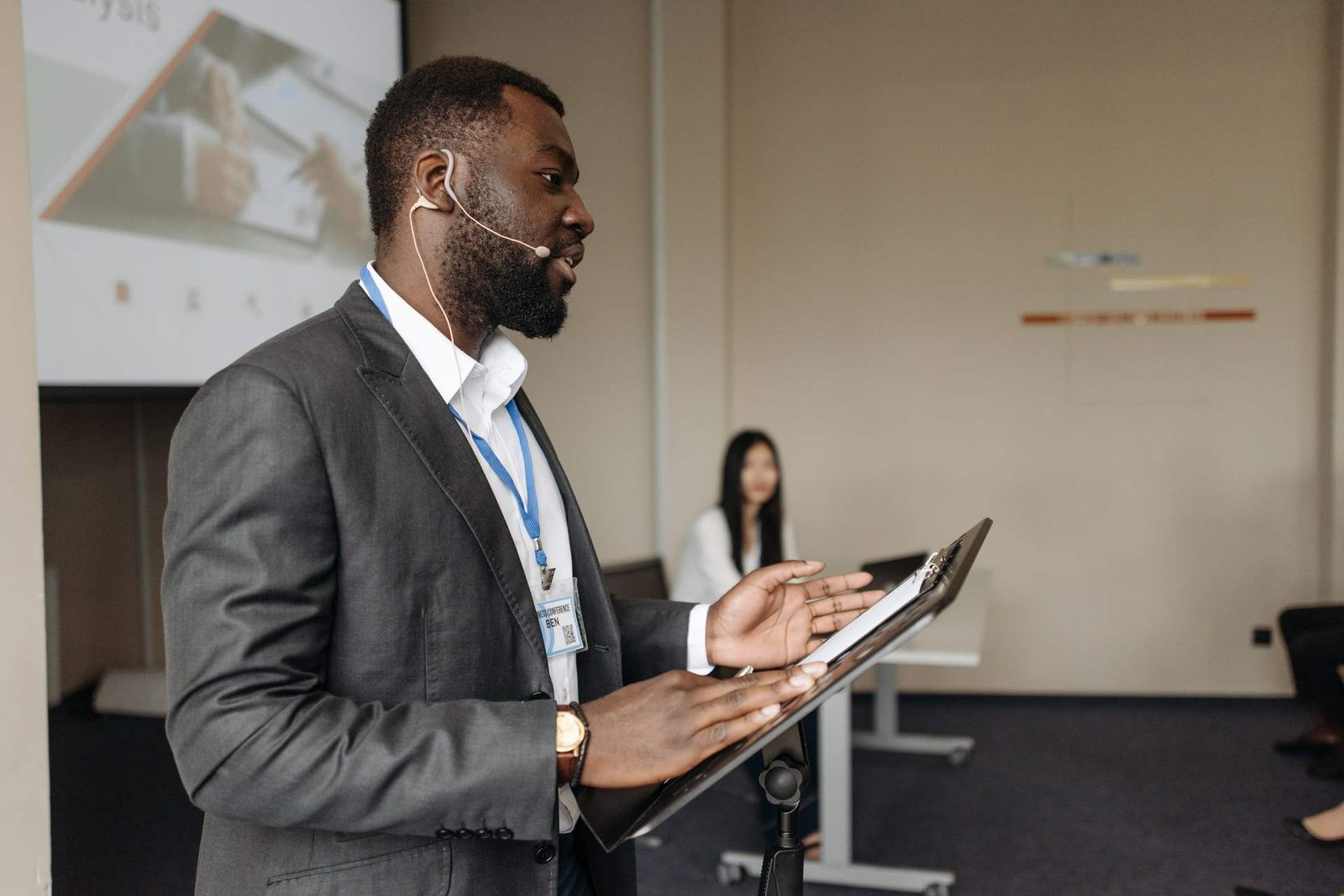 A presenter wearing a headset microphone speaks from a lectern during a business conference session, with a colleague seated behind