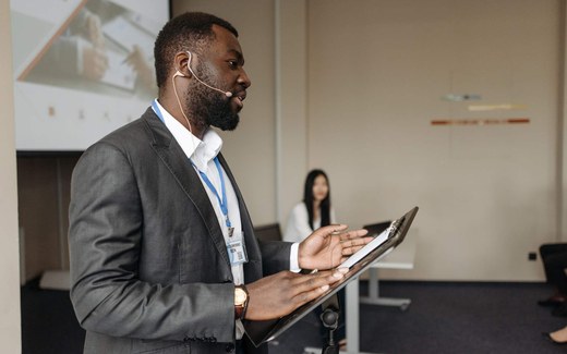 A presenter wearing a headset microphone speaks from a lectern during a business conference session, with a colleague seated behind