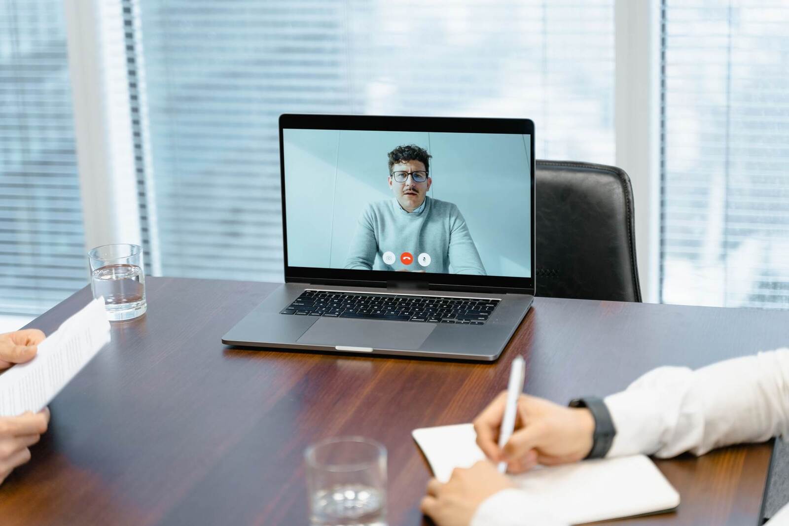 A laptop open on a boardroom table during a video call, with a notepad and pen in front of it