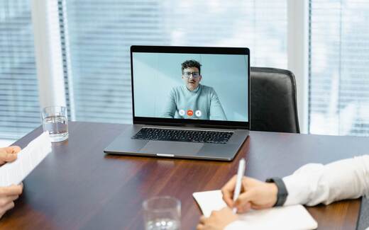 A laptop open on a boardroom table during a video call, with a notepad and pen in front of it