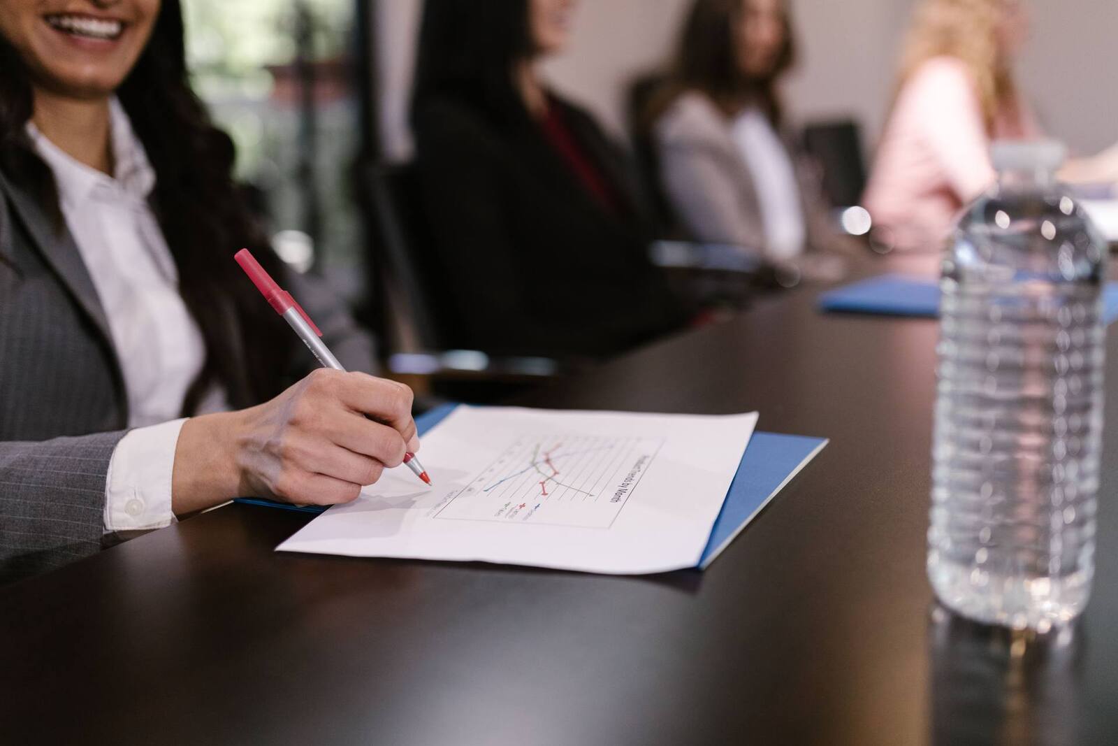 A smiling delegate in a pinstripe blazer marks a printed line chart with a red pen at a long meeting table, with blurred colleagues in business dress beyond