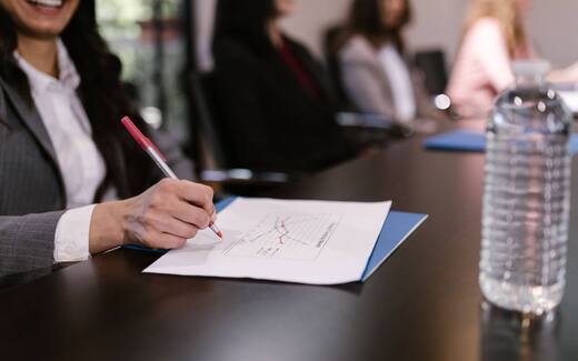A smiling delegate in a pinstripe blazer marks a printed line chart with a red pen at a long meeting table