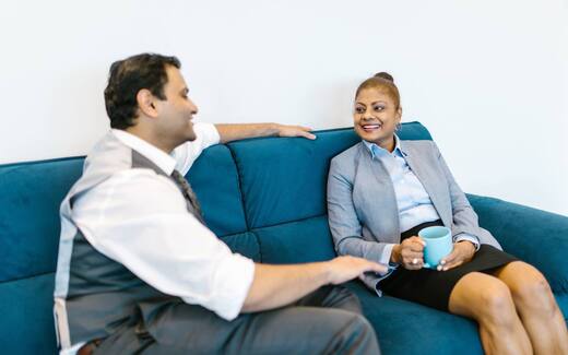 A woman in a grey blazer holds a mug and talks with a man in a waistcoat and tie on a teal sofa